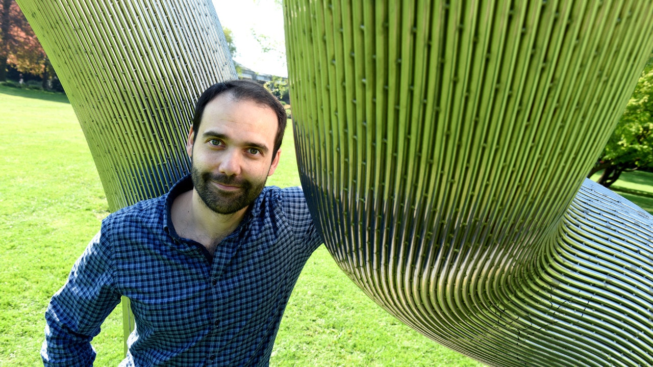 A man stands outside in a park next to a modern metal object.