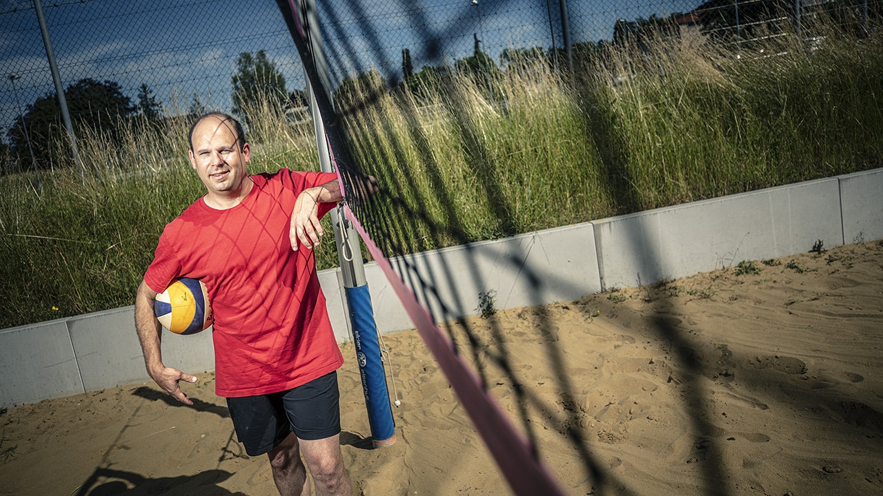 Man in red shirt and ball under his arm leaning against the beach volleyball net.