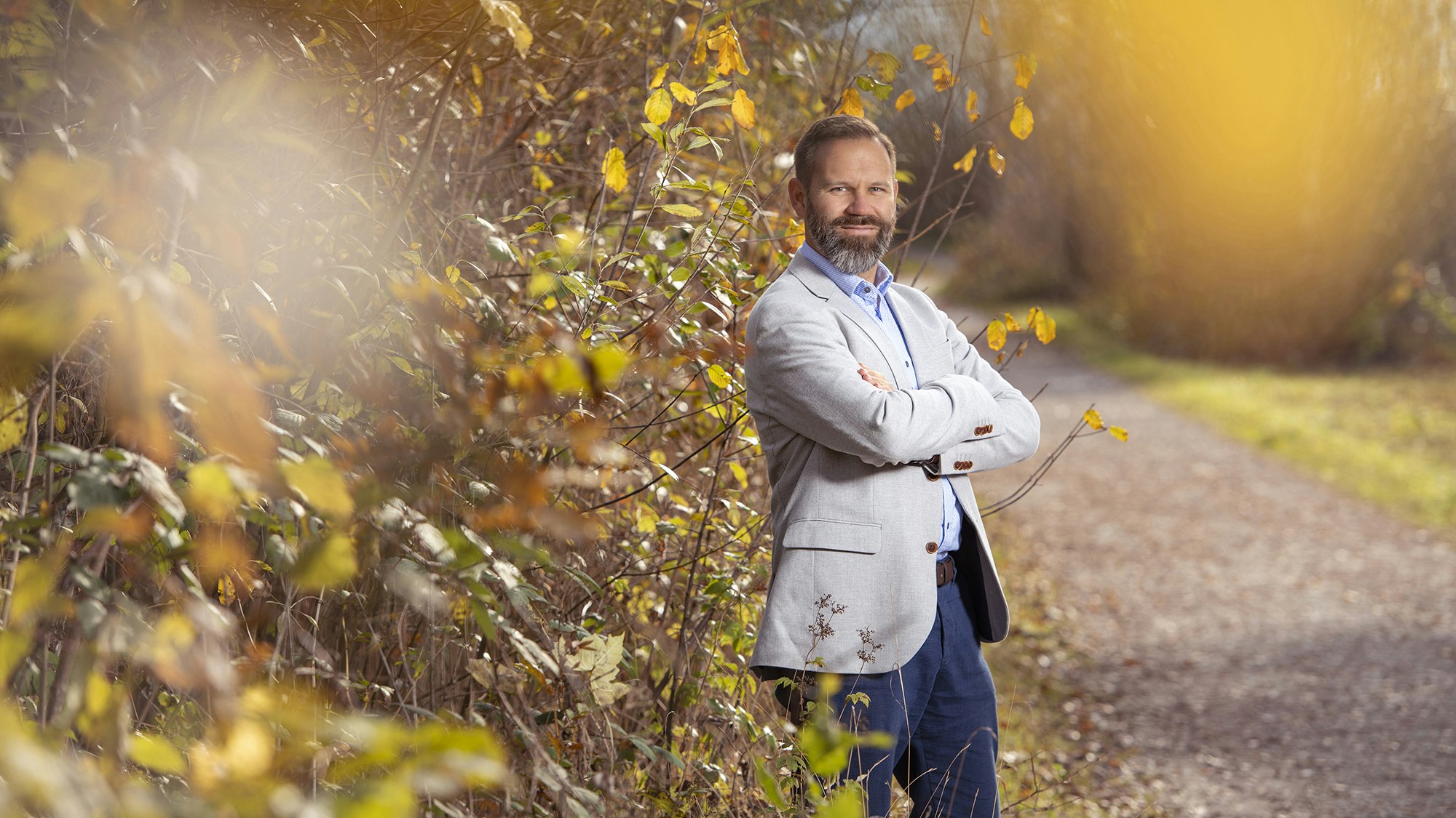 A man stands with folded arms in front of a golden bush.