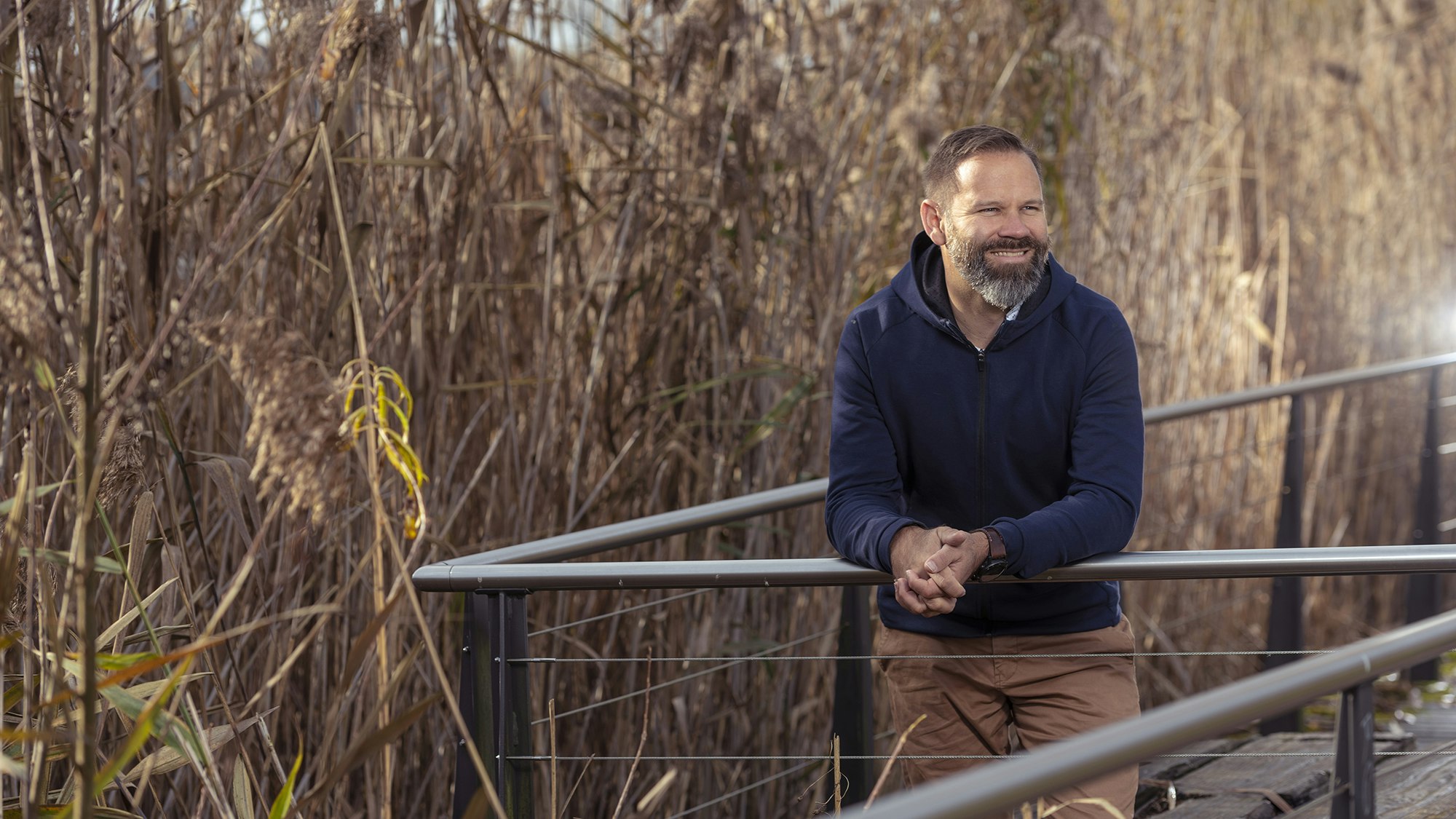 A man in casual clothes stands outside in front of a reed field.