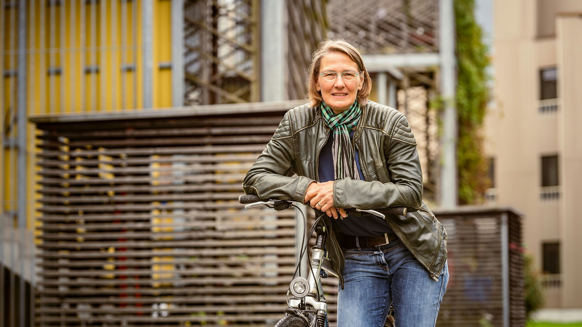 A woman in a leather jacket is standing with her bicycle in front of a residential building.