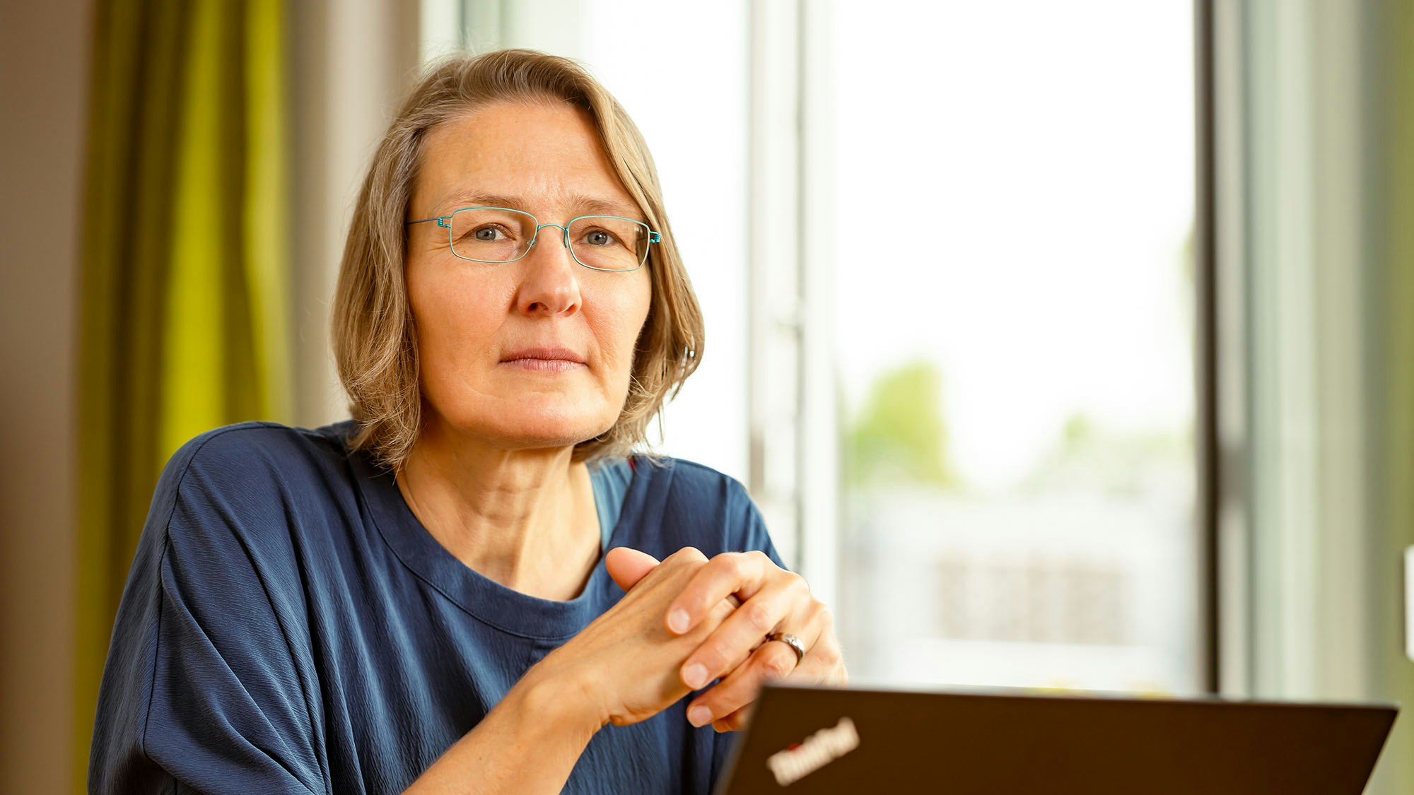 Woman with glasses sits at her PC with her hands clasped.