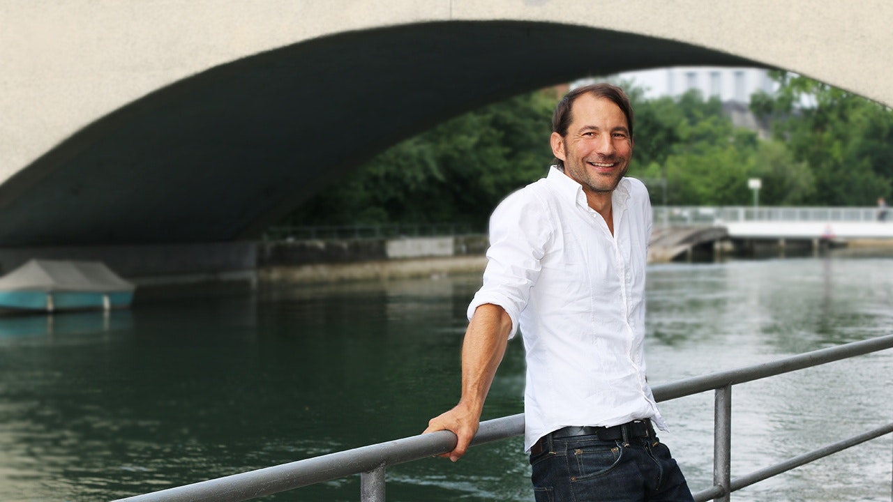 A man in a white shirt leans against the railing in front of a river.