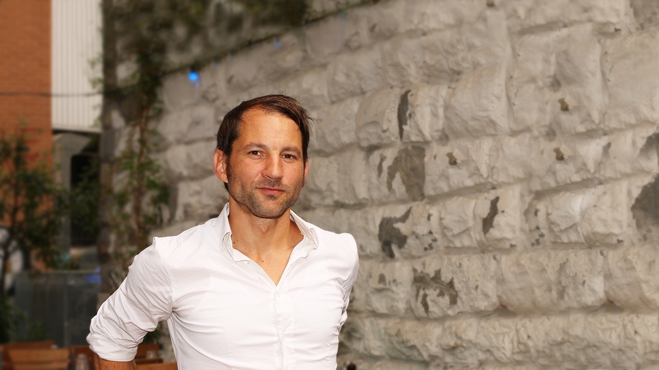 A man in a white shirt stands in front of a stone wall.