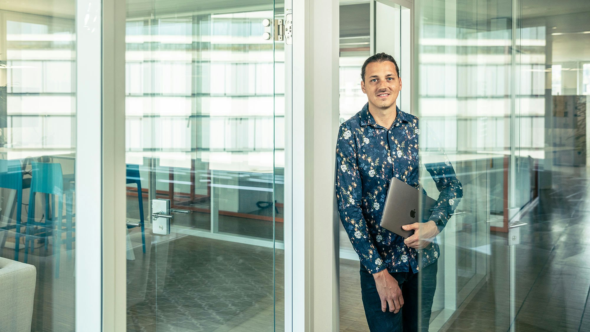 Man standing in the office with laptop in hand in the glass doorway.