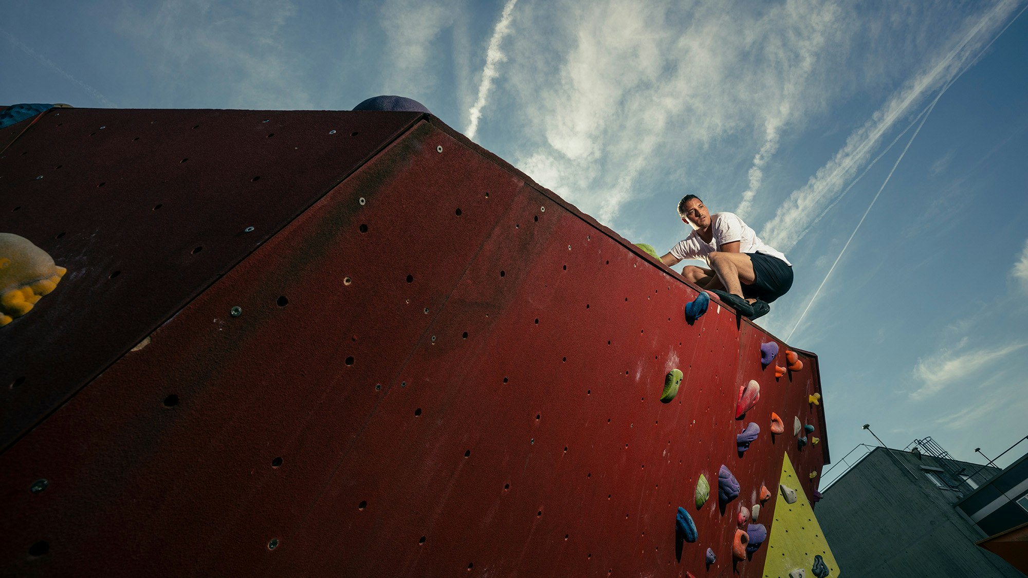 Mann sitzt ganz oben auf einer Kletterwand im Freien.