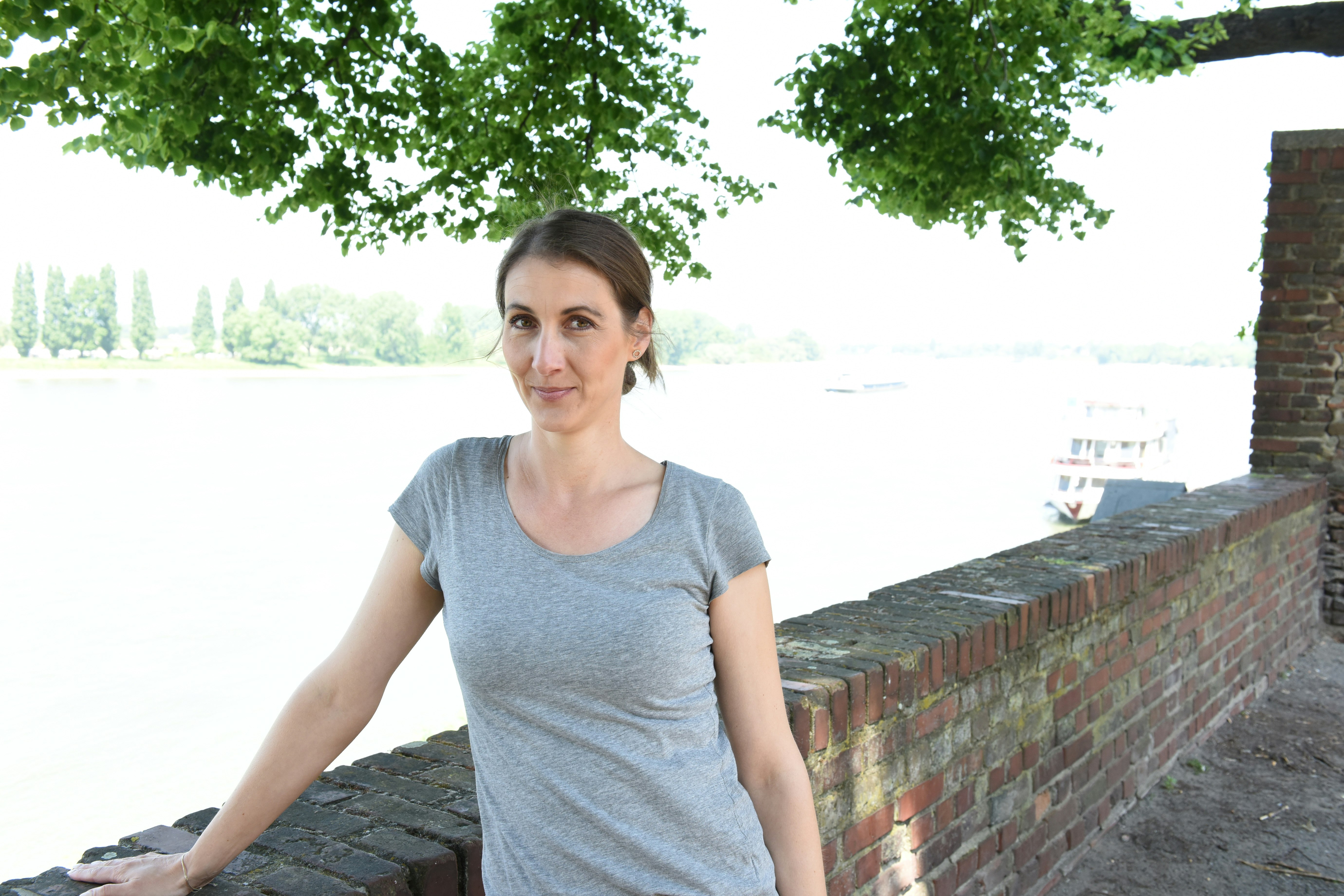 Katrin stands close to a brick wall wearing a grey t-shirt.