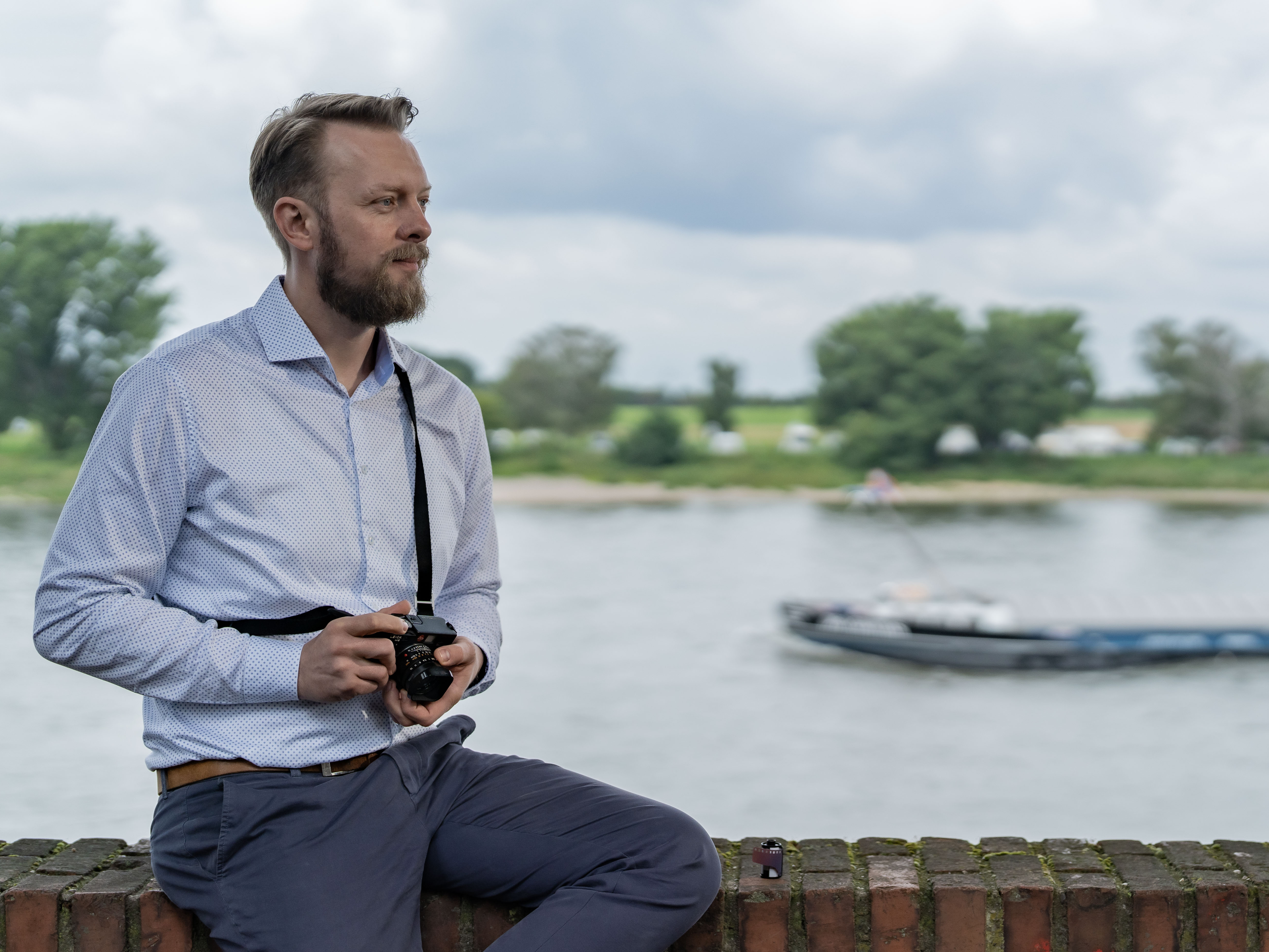 Damian sits on a brick wall at the river rhine looking to the right side while holding a camera.