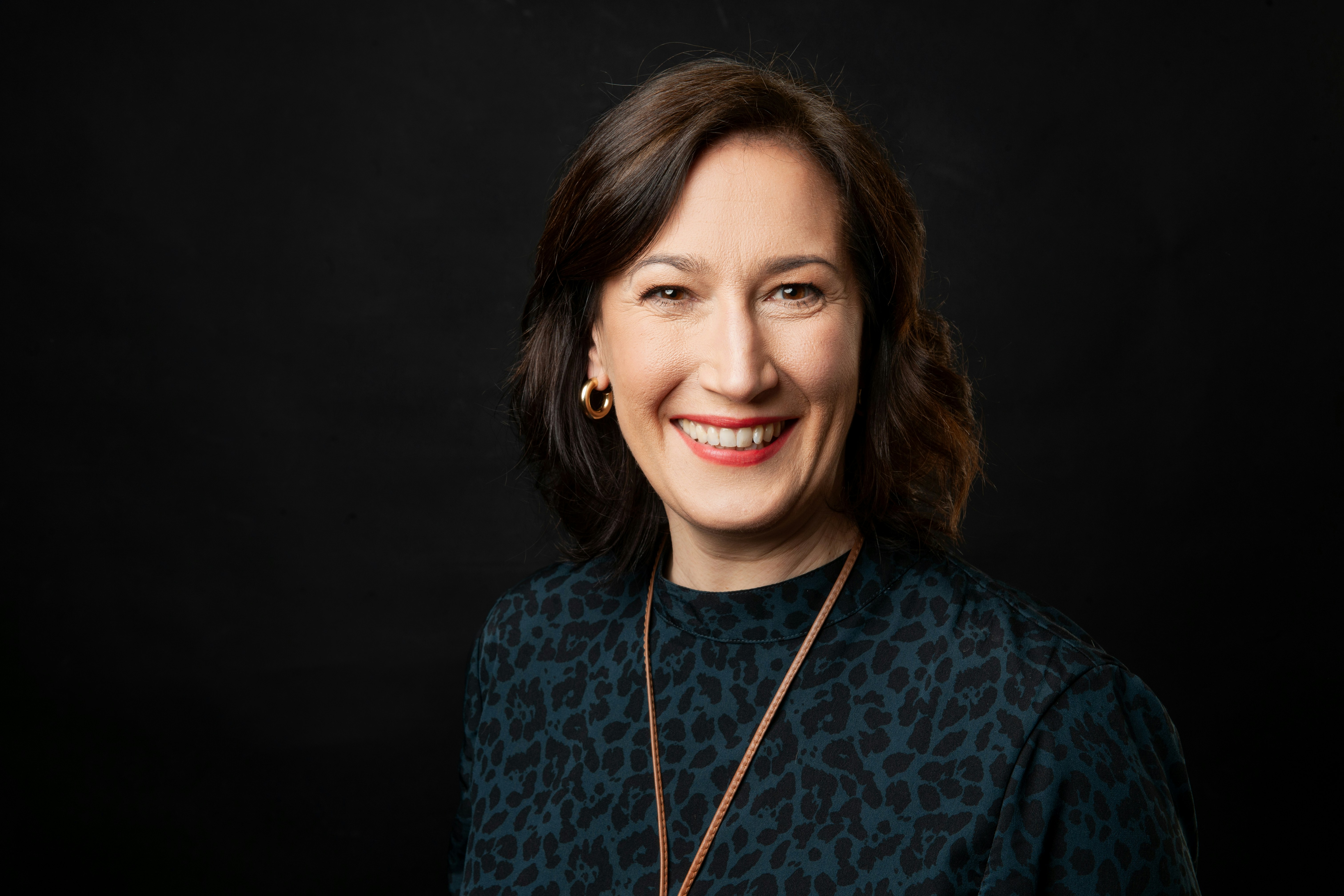 Julia smiles towards the camera wearing a necklace, standing in front of a black background.,