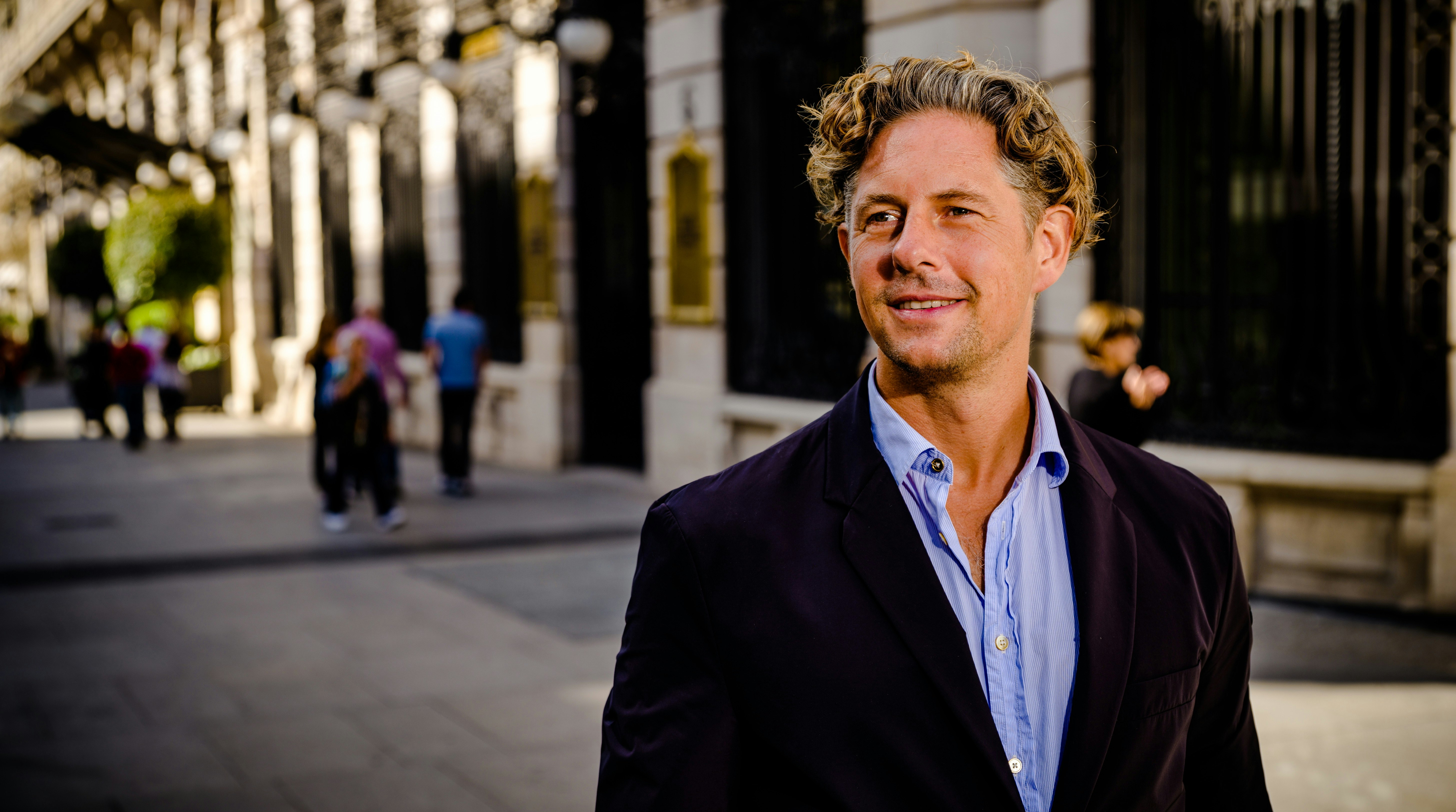Josef stands on the street, wearing a suit and smiles.