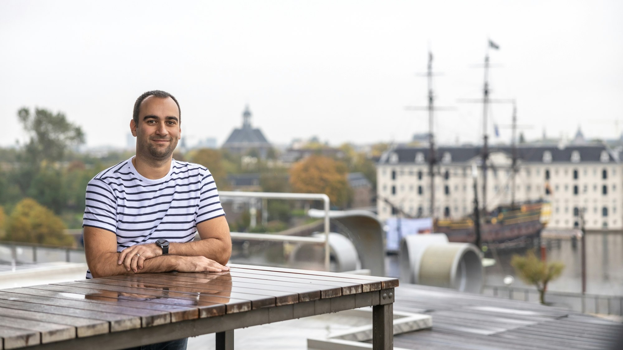 A man in a striped shirt on a terrace with Amsterdam in the background
