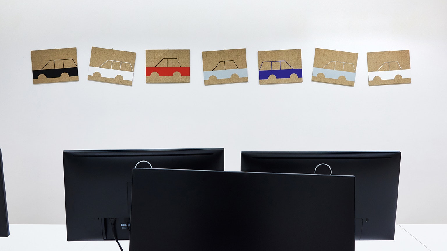 Im Vordergrund vier Monitore. An der Wand ein Werk aus einer Autokolonne, die nur aus bunten Karosserien besteht.