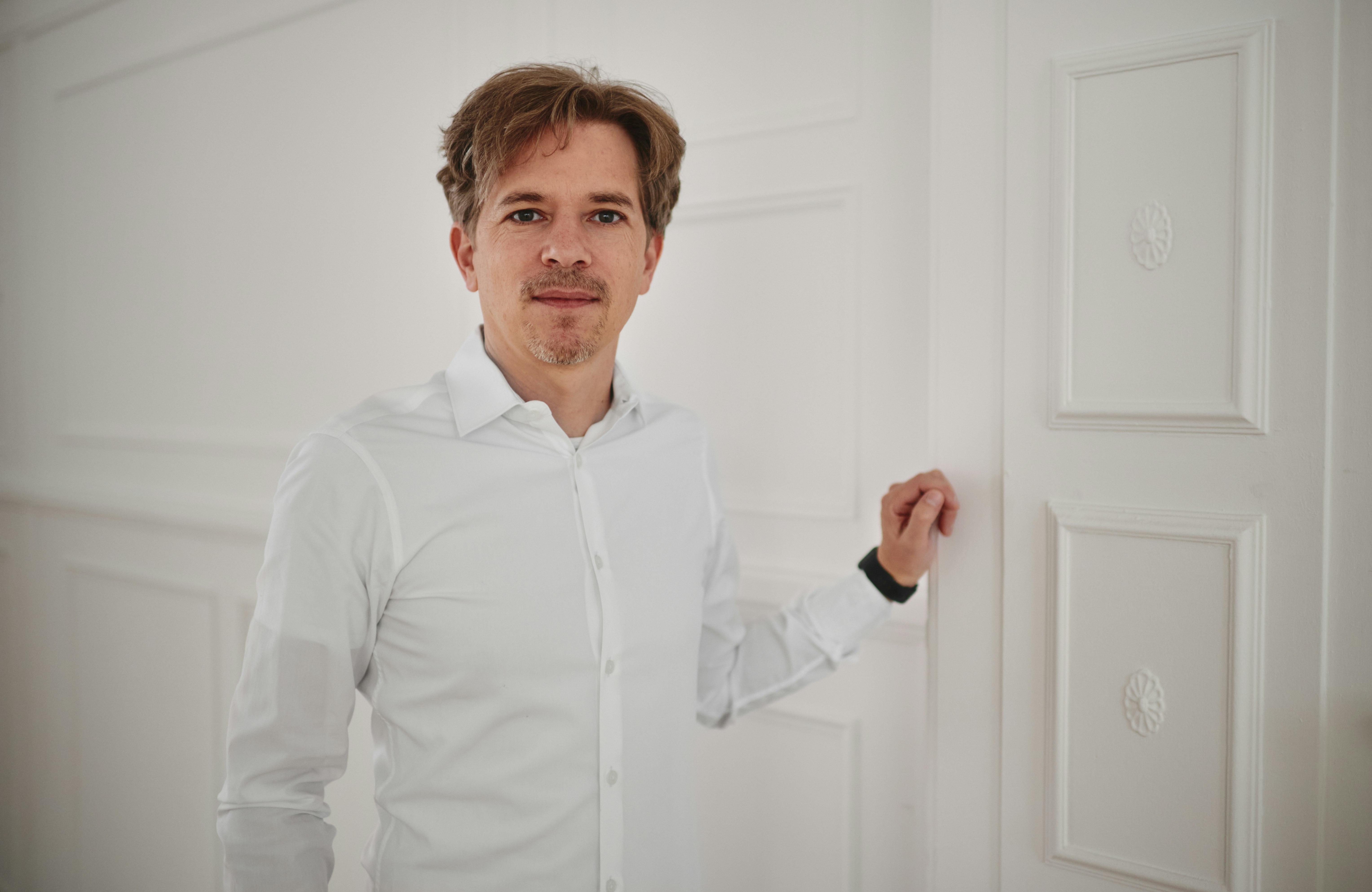 Christian Schröther lehnt an einer weißen mit Stuck verzierten Wand.