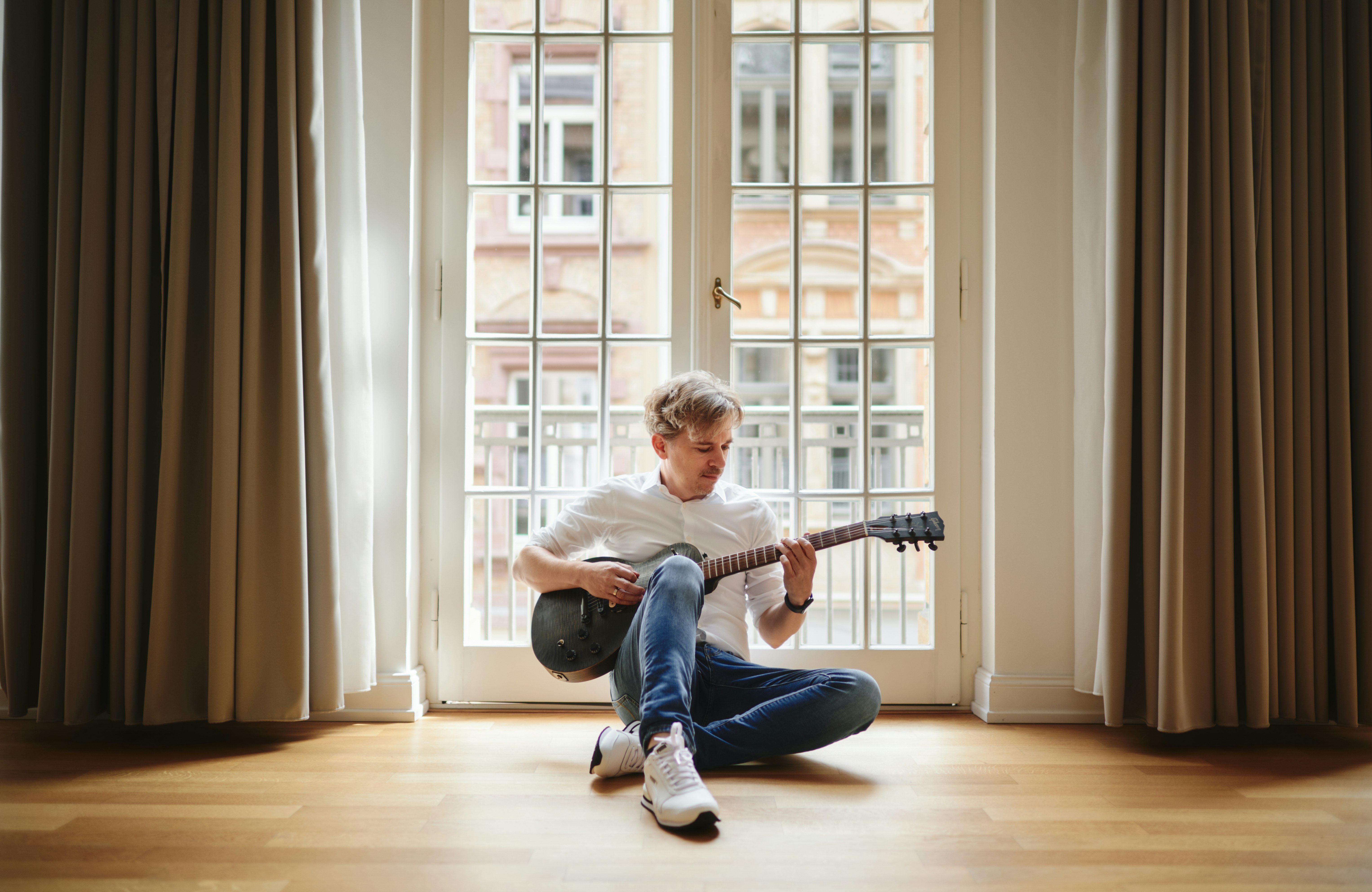 Christian Schröther sitzt mit einer Gitarre in der Hand vor dem Fenster auf dem Fußboden und spielt Gitarre