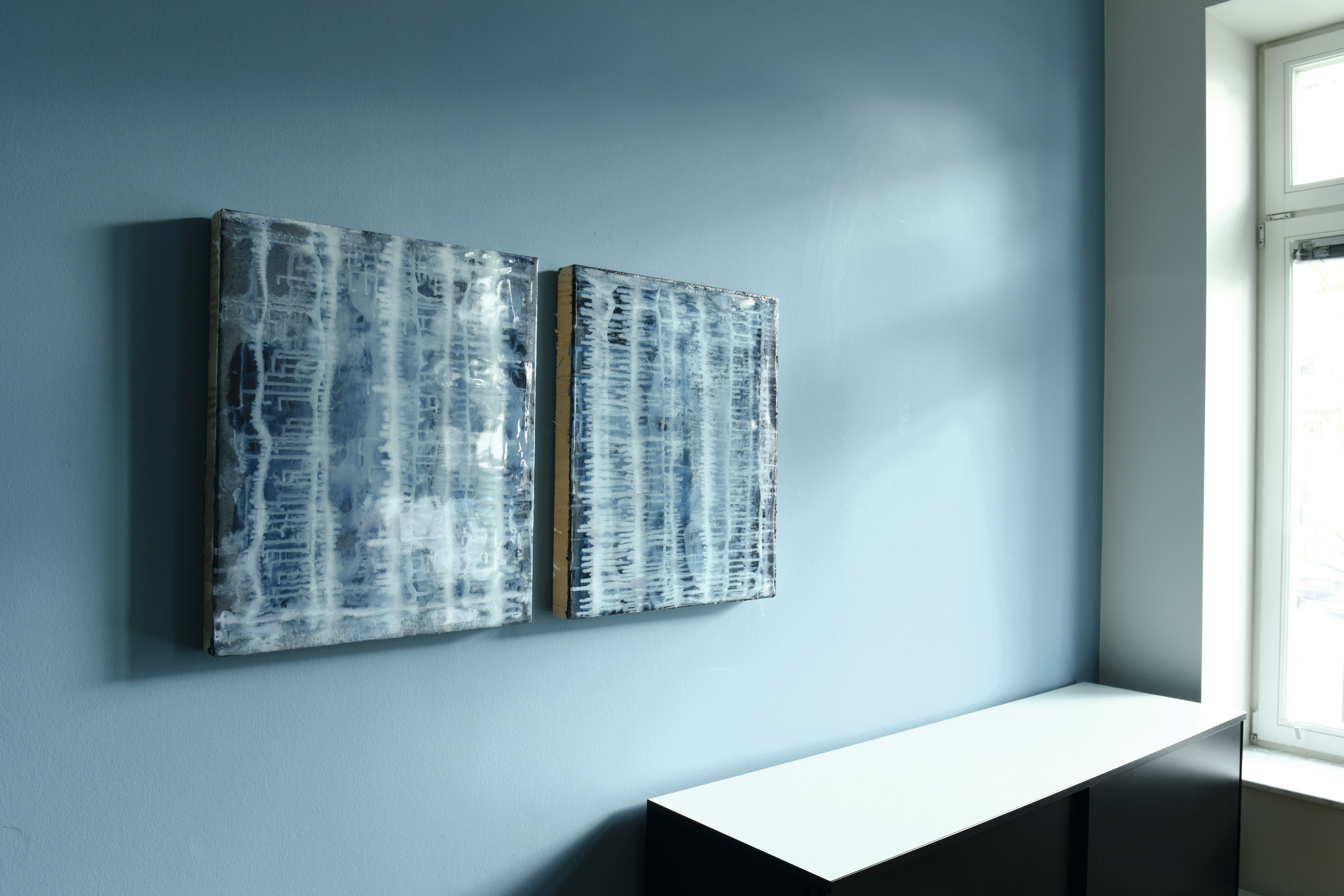 Zwei abstrakte, überwiegend blaue Gemälde mit vertikalen Linienstrukturen hängen nebeneinander an einer hellblauen Wand über einem weißen Sideboard.