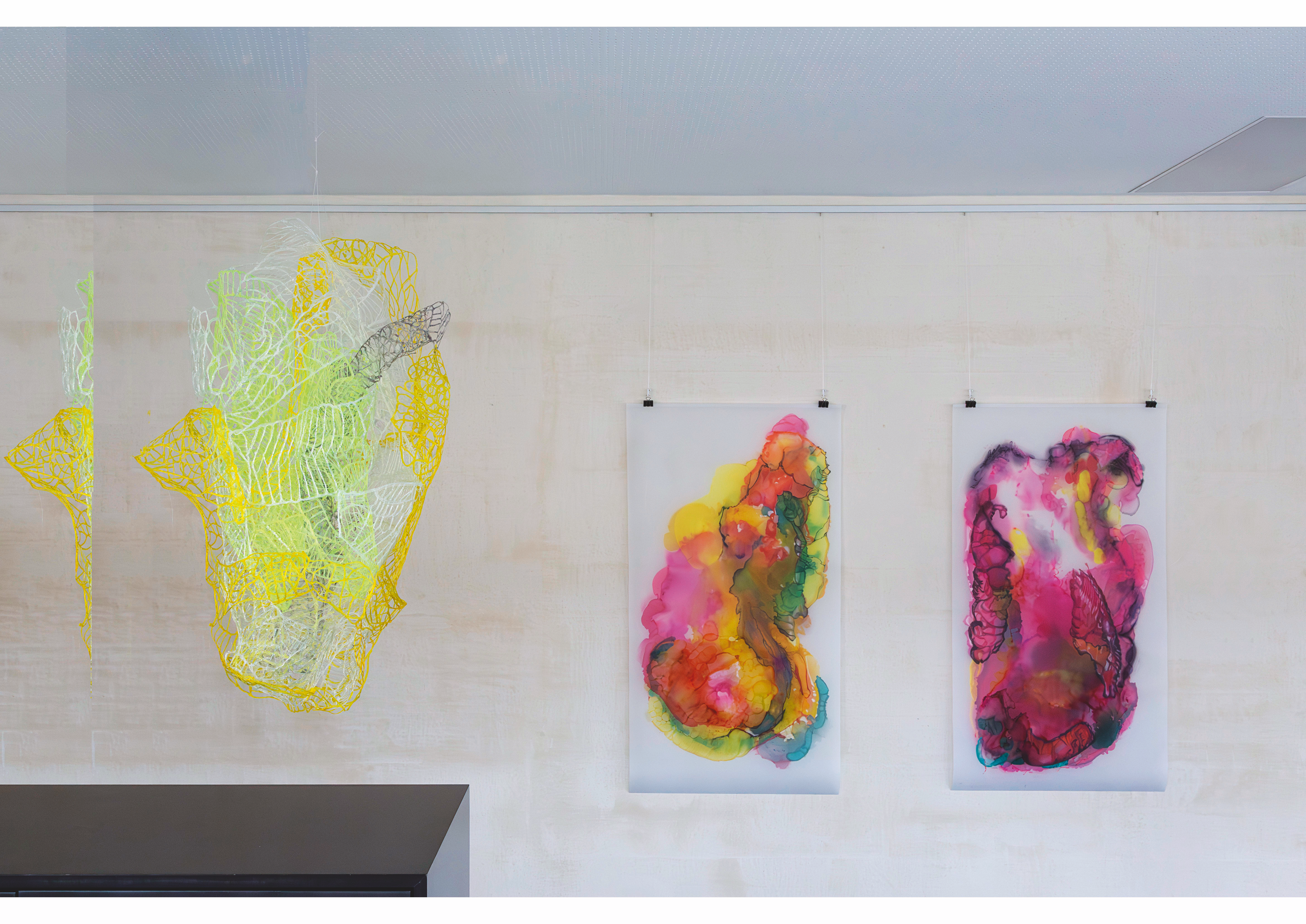 On a light wall, two colorful abstract artworks on translucent material hang side by side, center and right. To the left, a delicate wire sculpture in yellow, white, and grey with a mesh-like structure floats in the room."