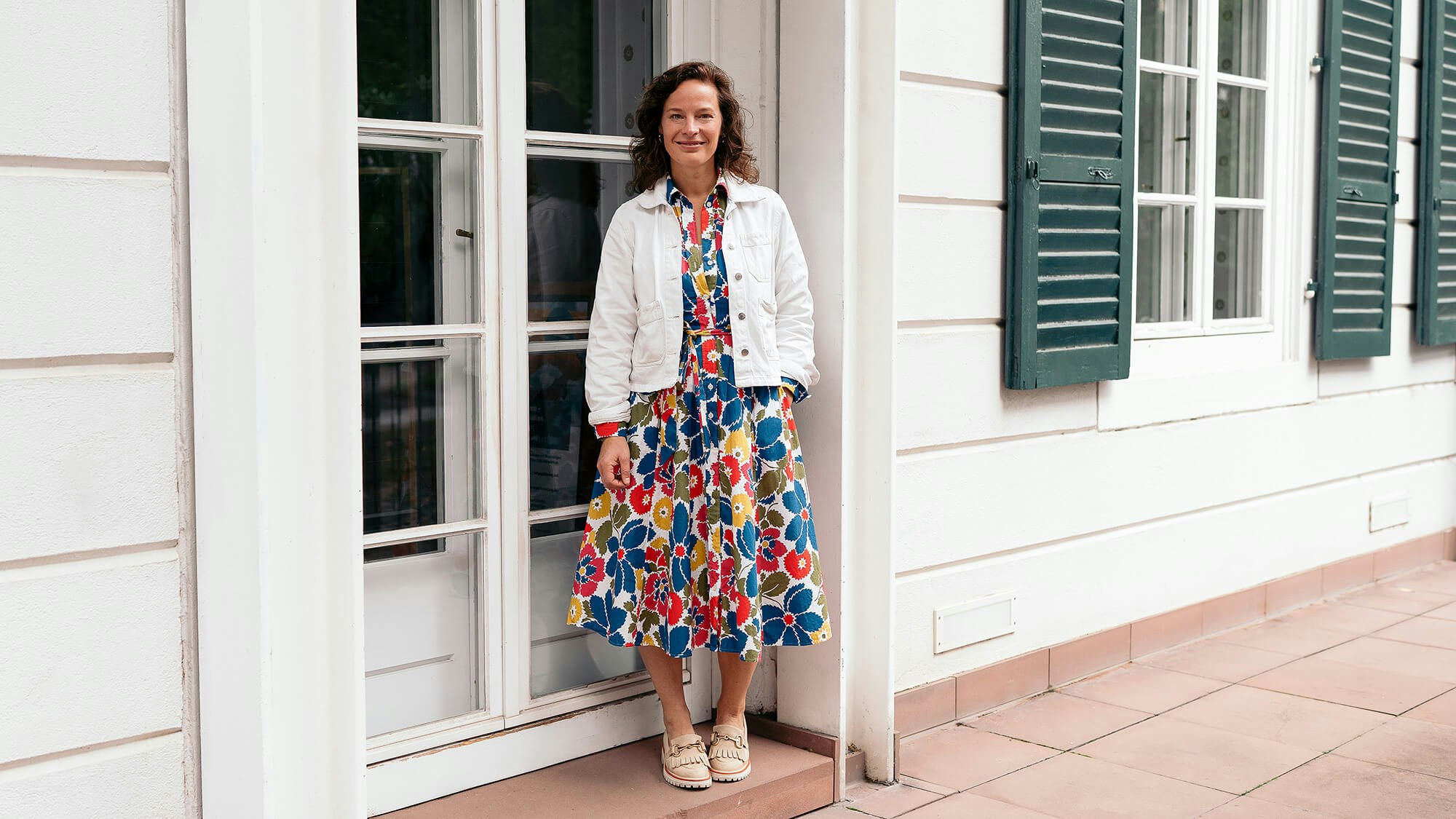 Juliane Huth steht vor einem hellen Gebäude mit Fensterläden und lächelt in die Kamera.