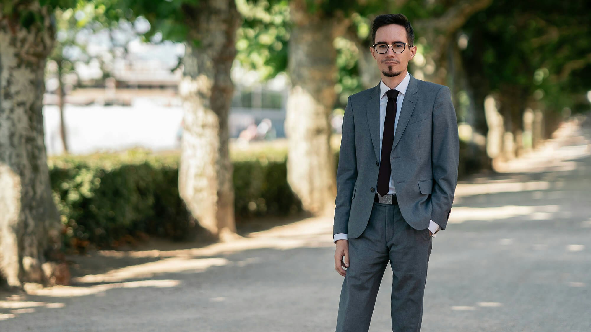 Christian Steinert in grauem Anzug steht auf einem von Bäumen gesäumten Weg im Freien und blickt in die Kamera.