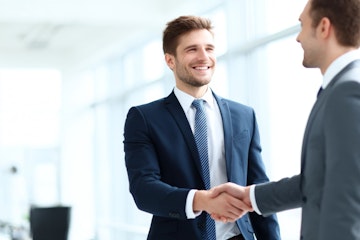 In a bright office, two business partners engage in a firm handshake, smiling and celebrating a successful meeting