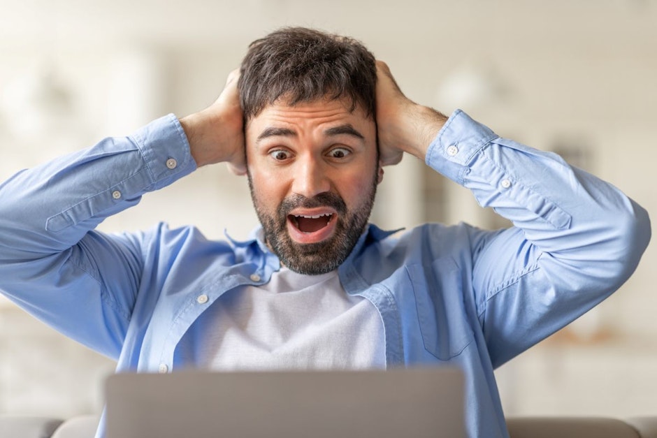 A man sits in front of a laptop, staring at the screen with shock and surprise