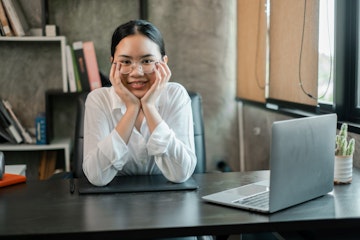 A young woman in a modern office, smiling confidently at her desk with a laptop and books, embodying professionalism and success.