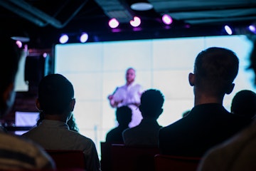 Point of View of people sat watching a speaker on stage who is slightly blurred.