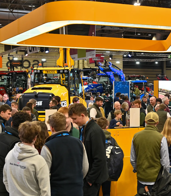 A group of people in the front wearing lanyards while in the background is a large yellow Tractor.