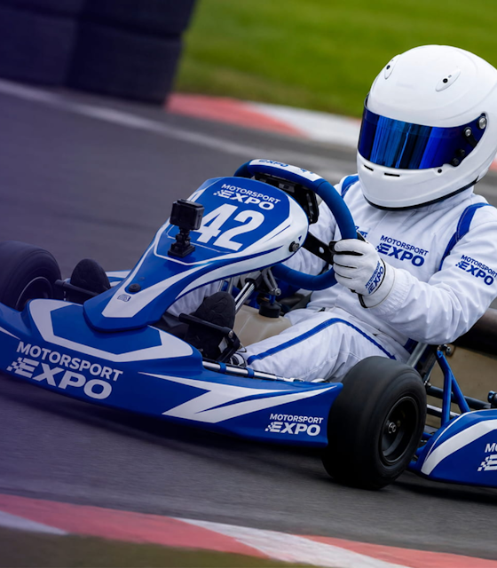 A boy dressed in racing gear driving a Go Kart on a track.