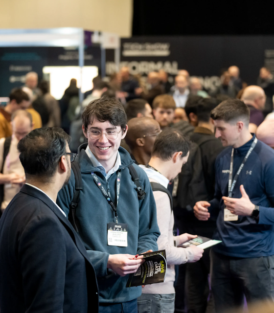 Two men talking to each other while in an exhibition hall surrounded by people, one of them is holding a brochure for the event.