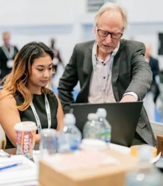 A man pointing towards a laptop which a young woman is using, likely instructing her.