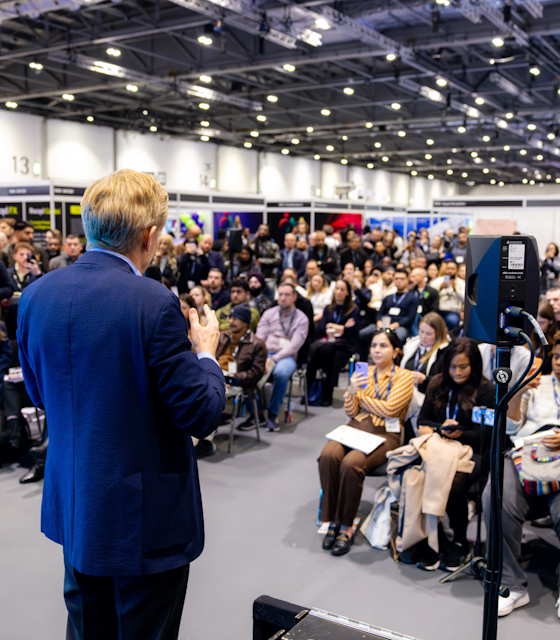 Speaker presenting to a large seated audience at Retrain Expo inside a busy exhibition hall.