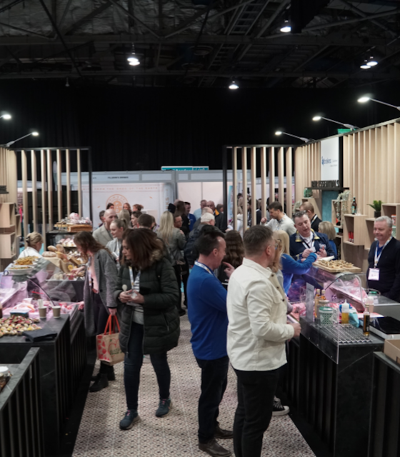 People stood in an exhibition stand looking at counters which are selling baked goods on either sides at ScotHot.