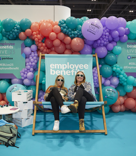 Two stylish women sat on a giant indoor blue deckchair with Employee Benefits written on it.