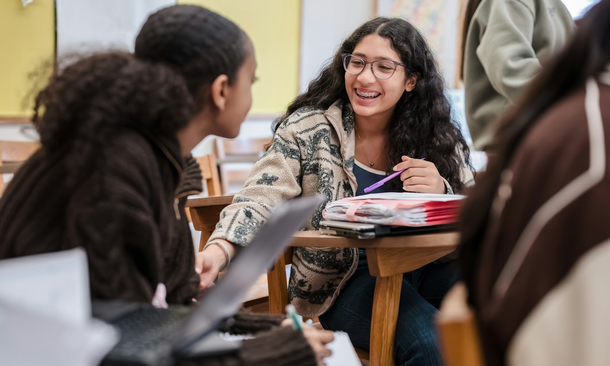 A smiling Upper School student at her desk covered in papers, pen in hand, gesturing to another student nearby