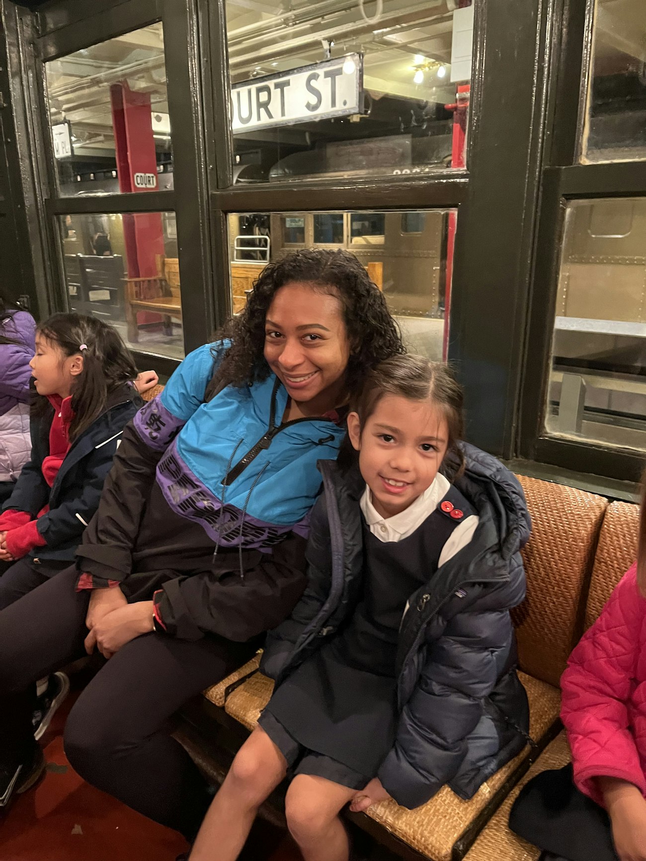 A lower school teacher and student smile and lean into each other sitting in a replica of an old New York subway car.