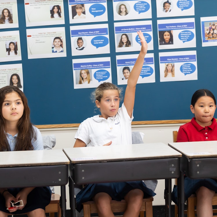 Three middle school students attentively listening to their teacher while one raises her hand eagerly.