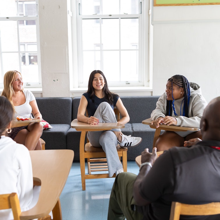 Upper school students sitting in a circle of desks smiling and engaging with each other.