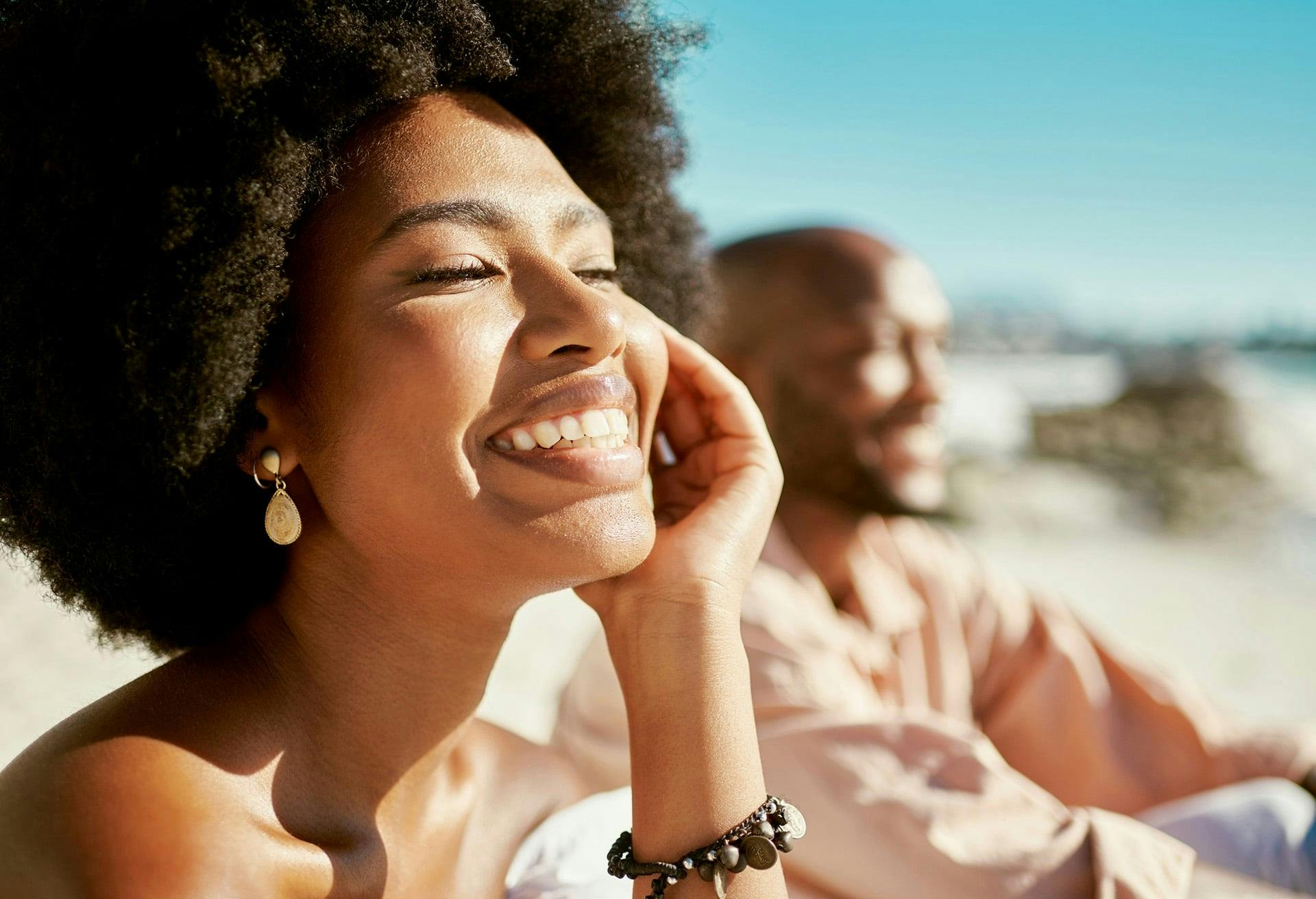 smiling woman with afro hair sitting on beach with man in background