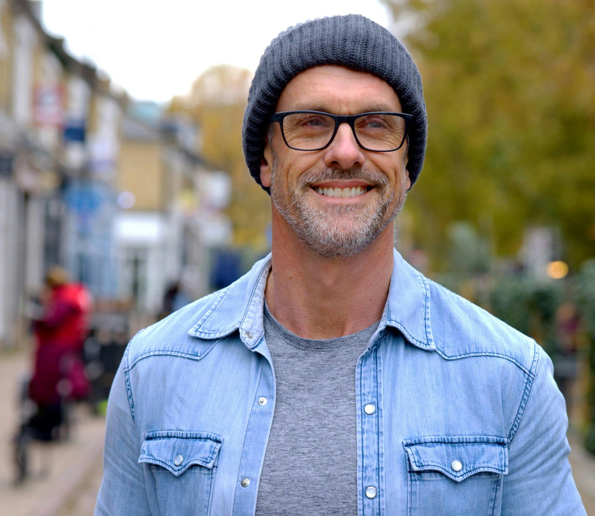 smiling man in a blue shirt and hat standing on a sidewalk