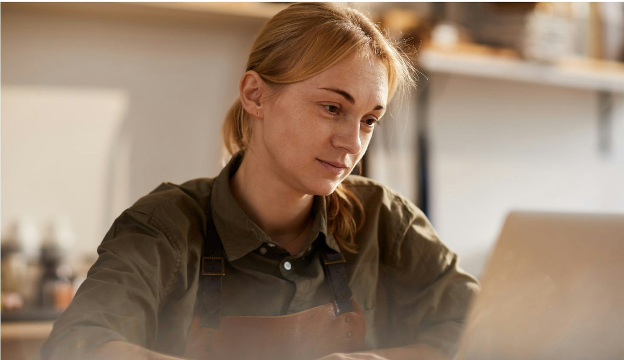 Woman in shirt and crafting apron working on laptop in workshop during sunset