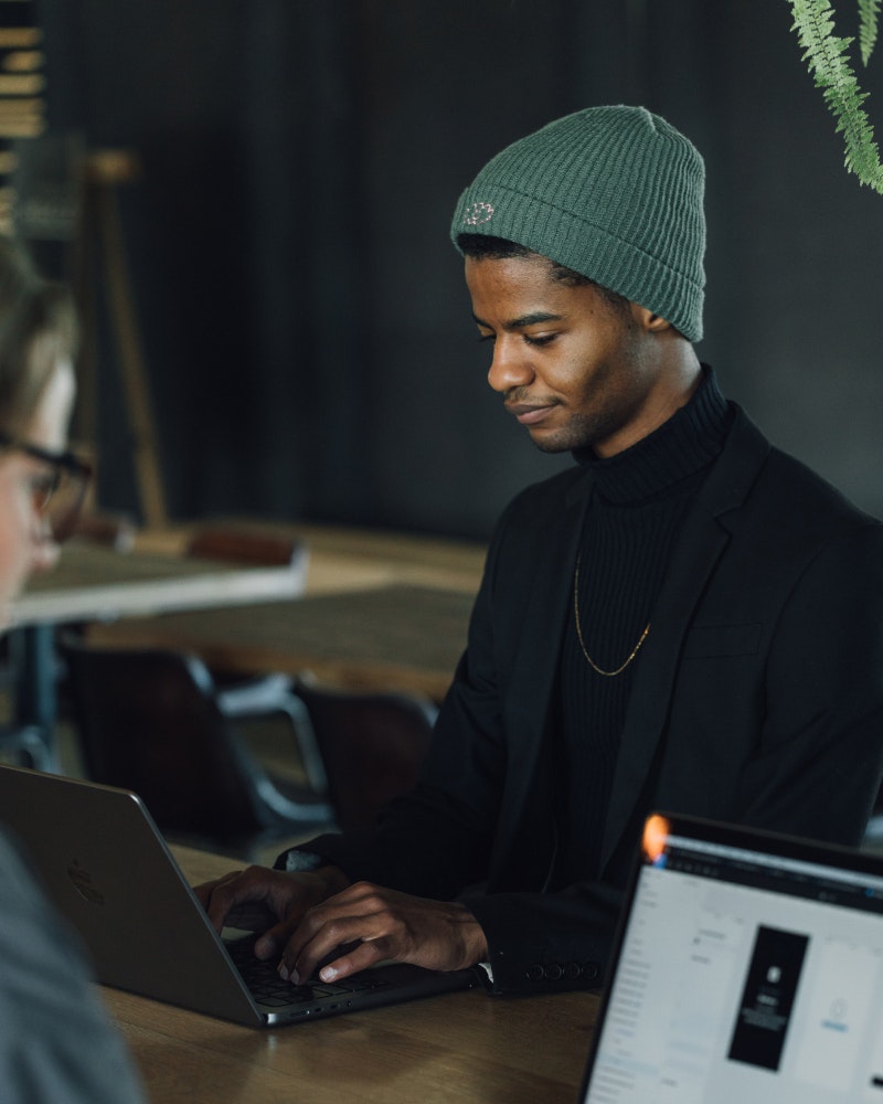 MakeReign worker sitting in office working Man Wearing a Beanie Working on MacBook in Office with Coworker on the Foreground