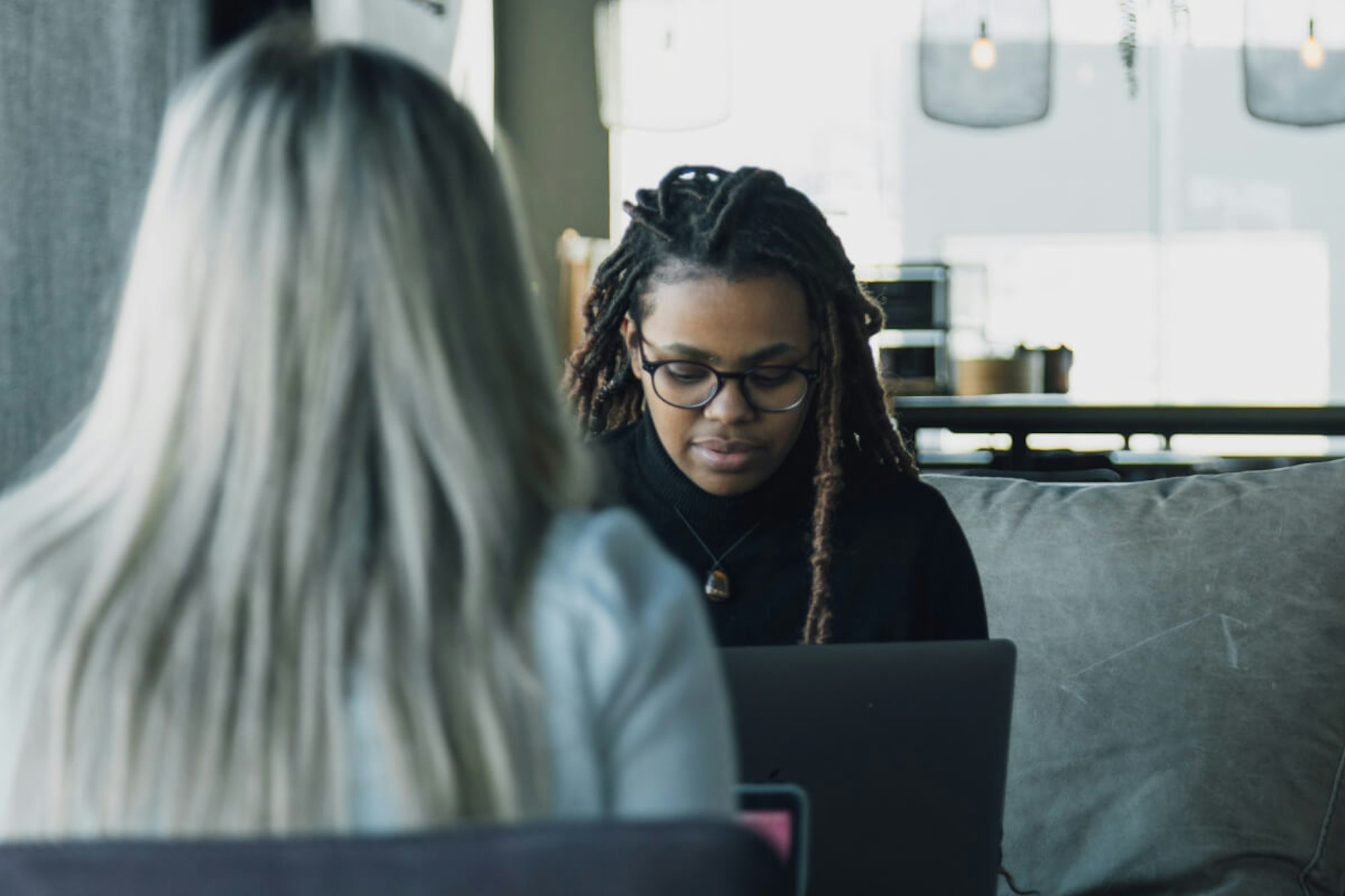 MakeReign women team members working at an office with laptops, one wearing glasses and locs in front view while blonde woman has her back to the camera