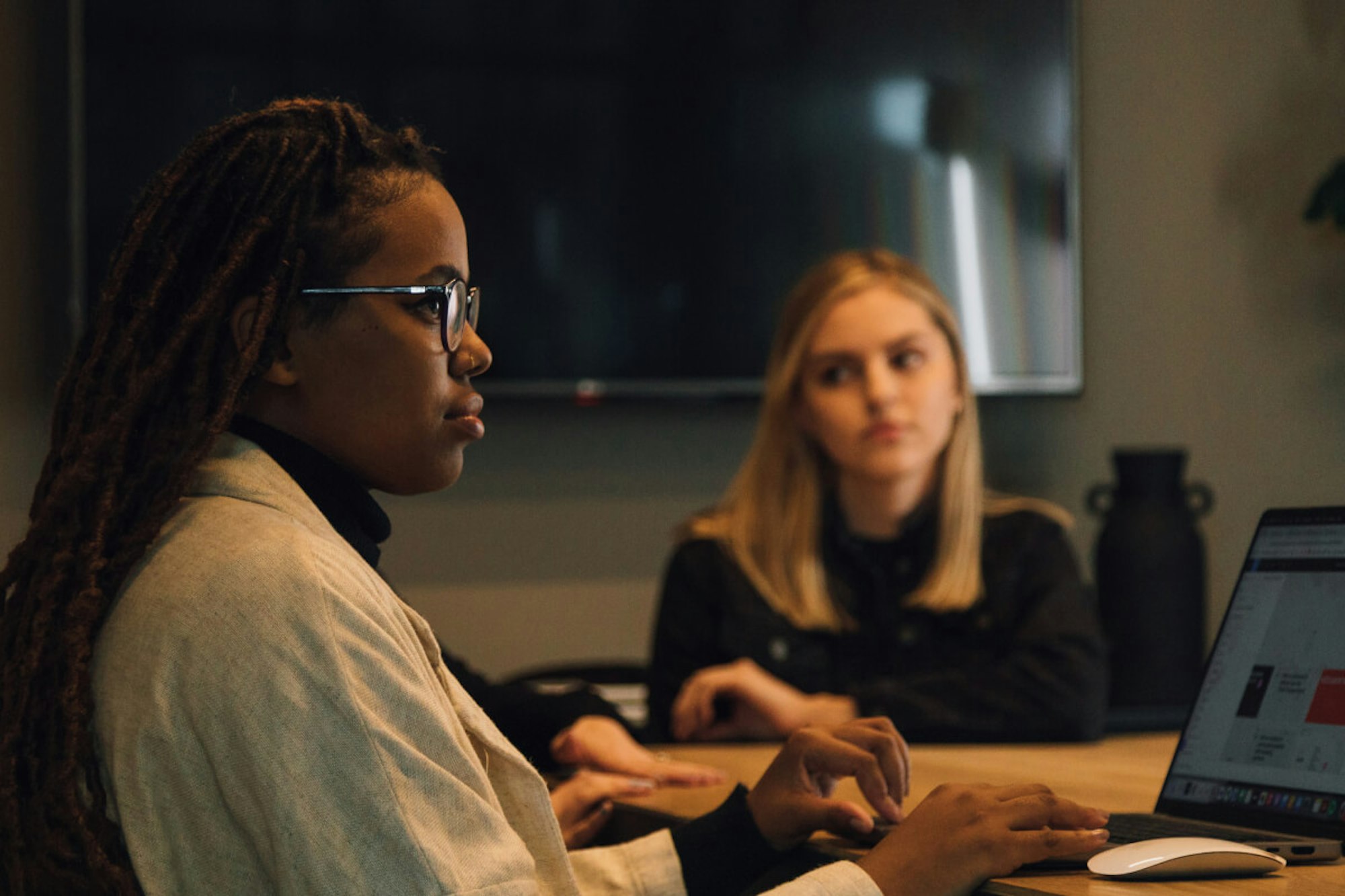 MakeReign women team members collaborating at an office with laptops, one wearing glasses and locs while blonde woman looks at the first woman with a screen in the background