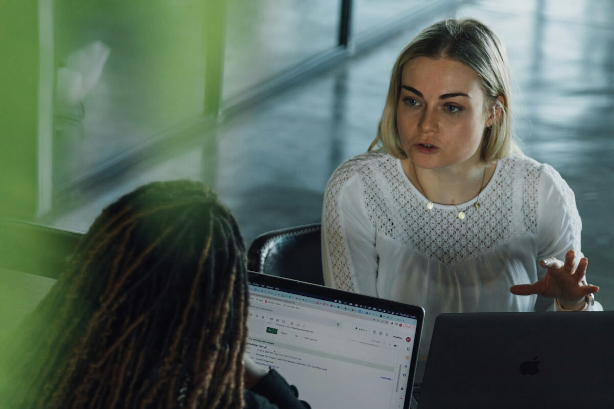 MakeReign women team members collaborating at an office with laptops, one wearing glasses and locs while blonde woman discusses with the first woman