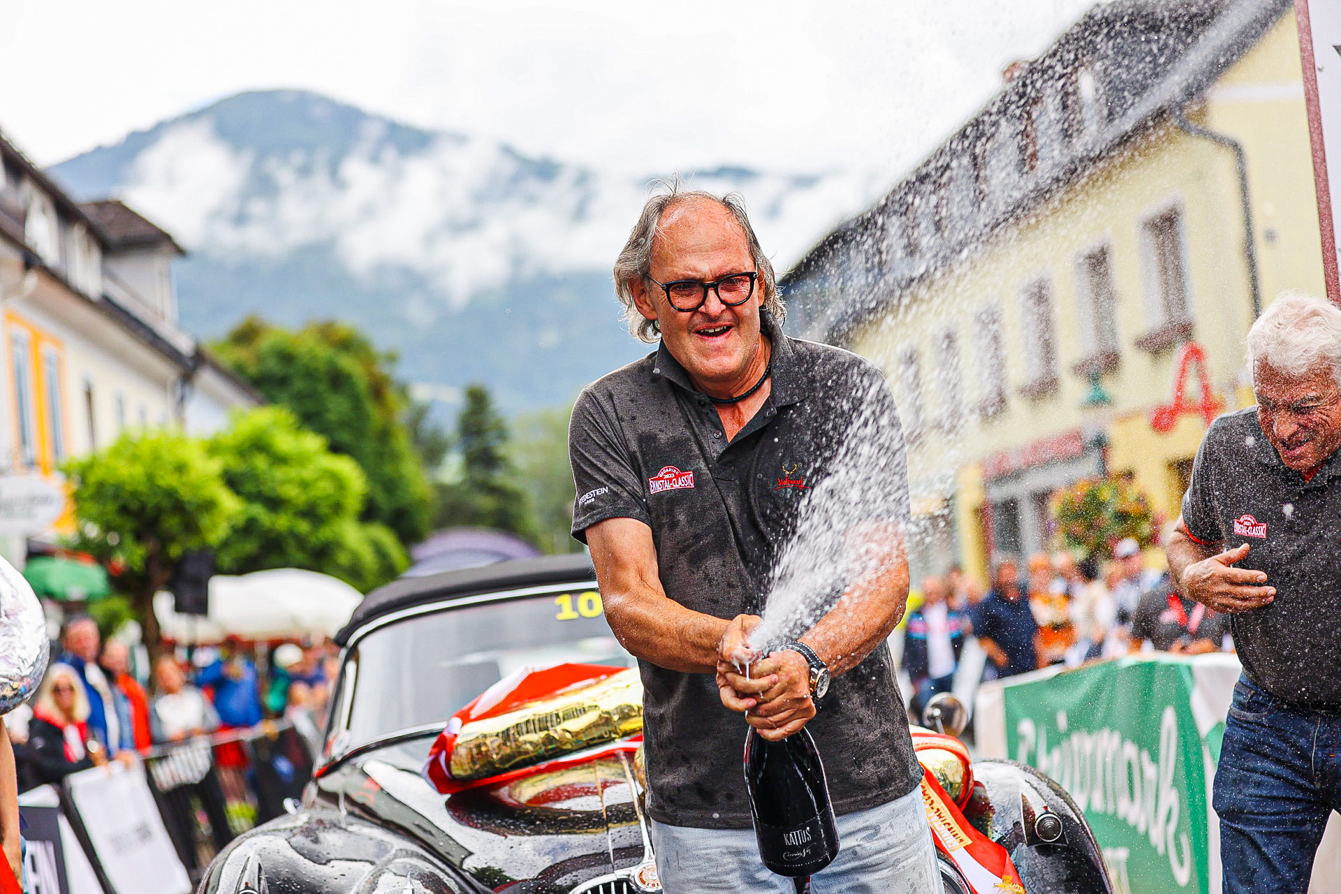Man celebrating with champagne in front of a classic car at the Ennstal-Classic event — BUBEN&ZORWEG
