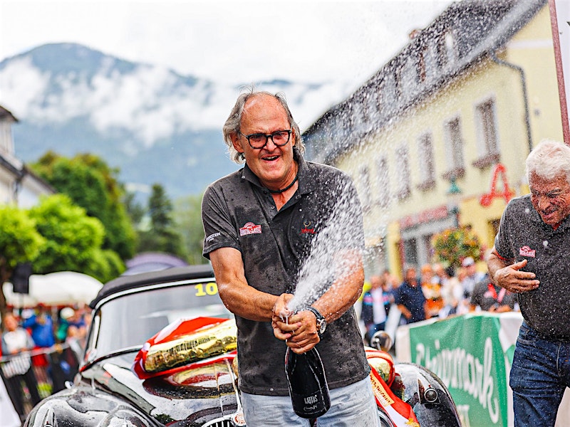 Man celebrating with champagne in front of a classic car at the Ennstal-Classic event — BUBEN&ZORWEG