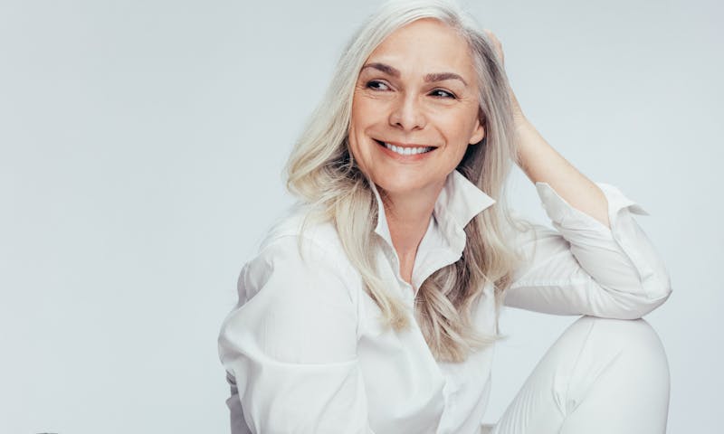 Older woman wiht long gray hair smiles looking away from the camera. Her elbow rests on her knee. Her youthful appreance despite her age, is to show the potential results of a brow lift with Dr. Moradi.