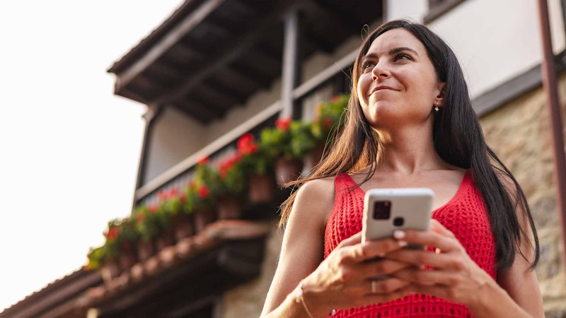 Woman looking into the distance holding her phone while standing infront of here house.