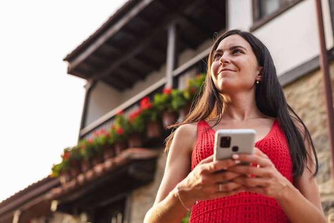 Woman looking into the distance holding her phone while standing infront of here house.