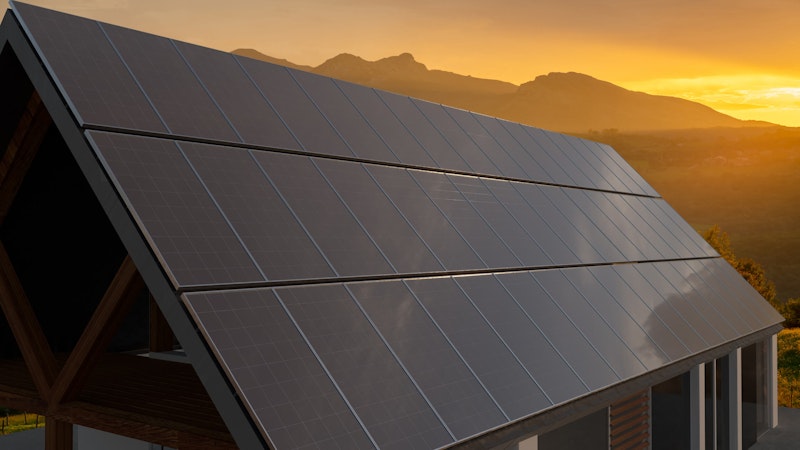 Black solar panels set on a steap angled roof backlit by the sunest over the mountains